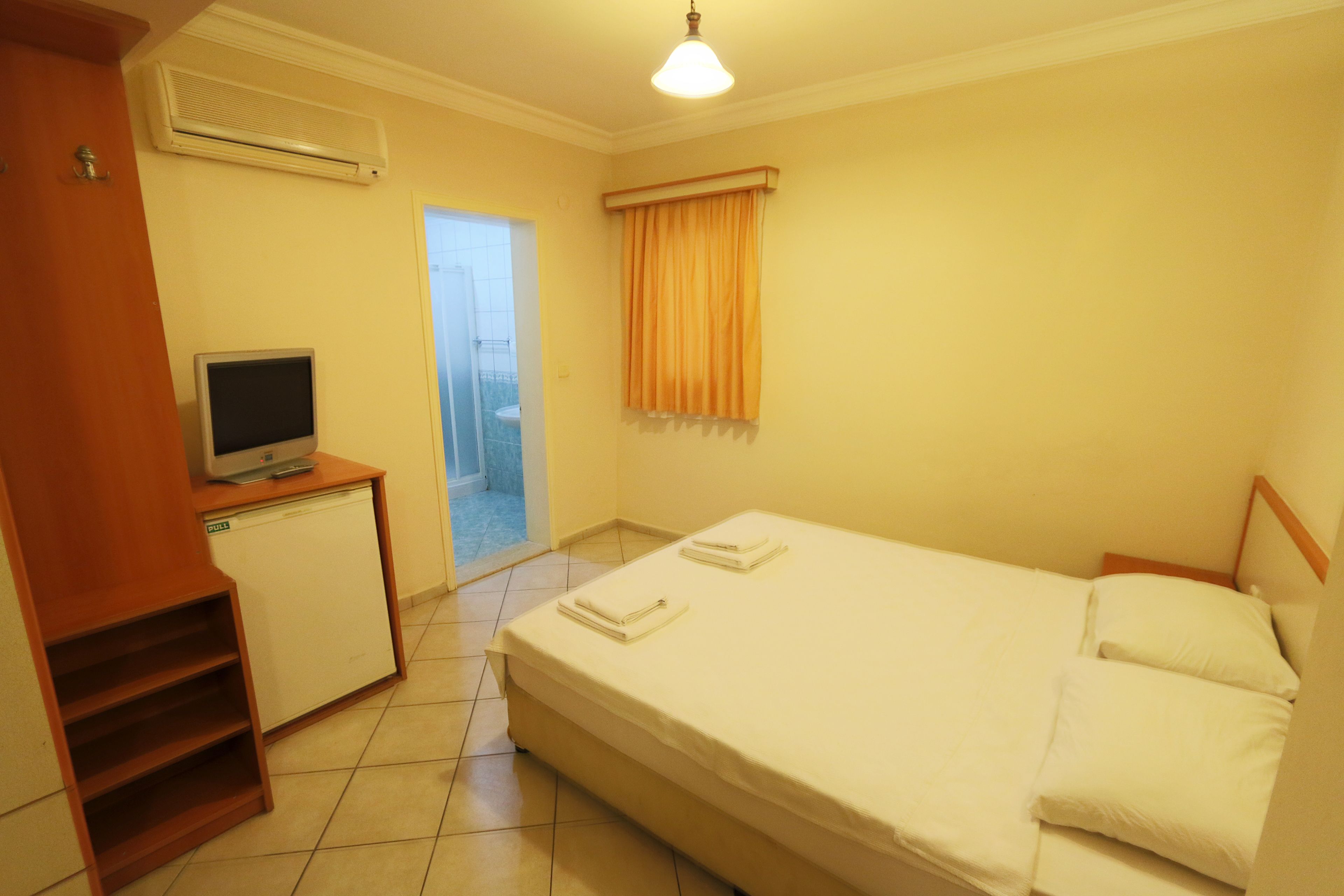 Una camera d'albergo modesta con un letto, una scrivania con un computer, un piccolo frigorifero e una finestra con tende.