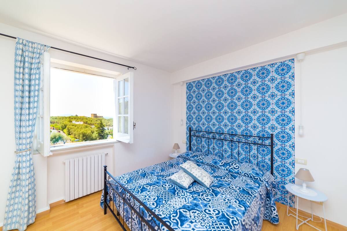 Une chambre avec une couvre-lit bleue et blanche à motifs, une vue sur la verdure par la fenêtre et un radiateur blanc.