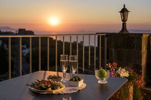 Una mesa con dos vasos de agua, un cuenco de aceitunas y un bol de fruta en un balcón con vistas a la puesta de sol.