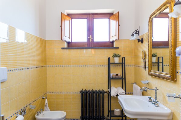 Un baño con paredes de azulejos amarillos, una ventana con persianas abiertas, un radiador, un lavabo, un inodoro y un estante con artículos de aseo.