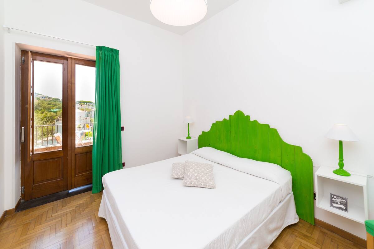 Une chambre avec une porte en bois, des rideaux verts, un lit avec une literie blanche et une tête de lit verte.