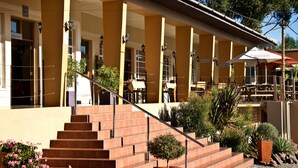 A building with a covered outdoor seating area, a staircase leading to the entrance, and various potted plants. - Bahnhof Hotel Aus (Aus)