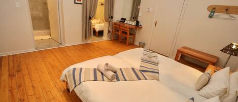 Une chambre avec un plancher en bois, un lit avec une literie à rayures, un bureau avec une chaise, un miroir et une porte donnant sur une salle de bain.