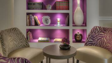 A modern living room with a purple-lit bookshelf, two beige armchairs, and a round coffee table.