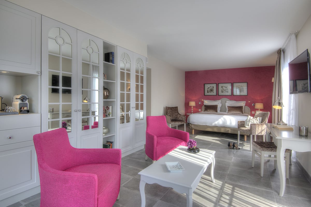 Et hotelværelse med en seng, to pink farvede stole, et hvidt bord og et hvidt skrivebord.