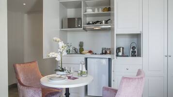 Une cuisine compacte avec des meubles blancs, un four à micro-ondes, un réfrigérateur et un petit plan de travail. On y trouve deux chaises roses disposées autour d’une table blanche, sur laquelle se trouve un vase de fleurs.
