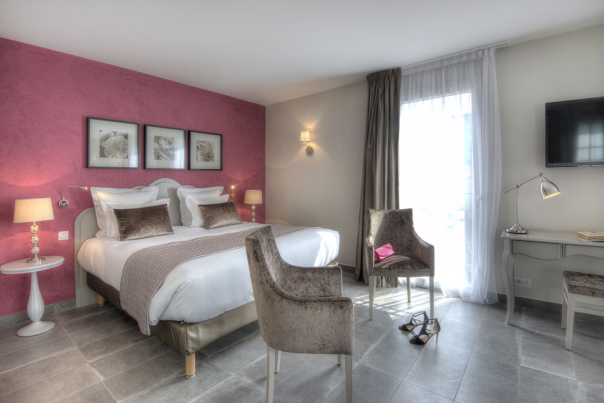 Et hotelværelse med en seng, to stole, et skrivebord, et fjernsyn og et vindue med gardiner.