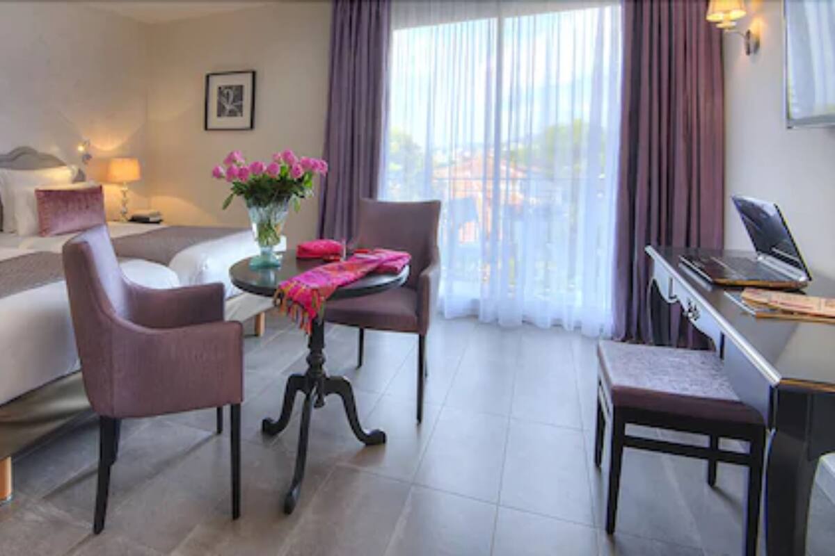 Et hotelværelse med en seng, to stole, et skrivebord med en bærbar computer, en vase med blomster og et vindue med gardiner.
