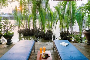A serene outdoor seating area with two lounge chairs, a small table with a book and drinks, surrounded by lush greenery and a calm water body. - Riverside Oasis Villa (Da Nang)