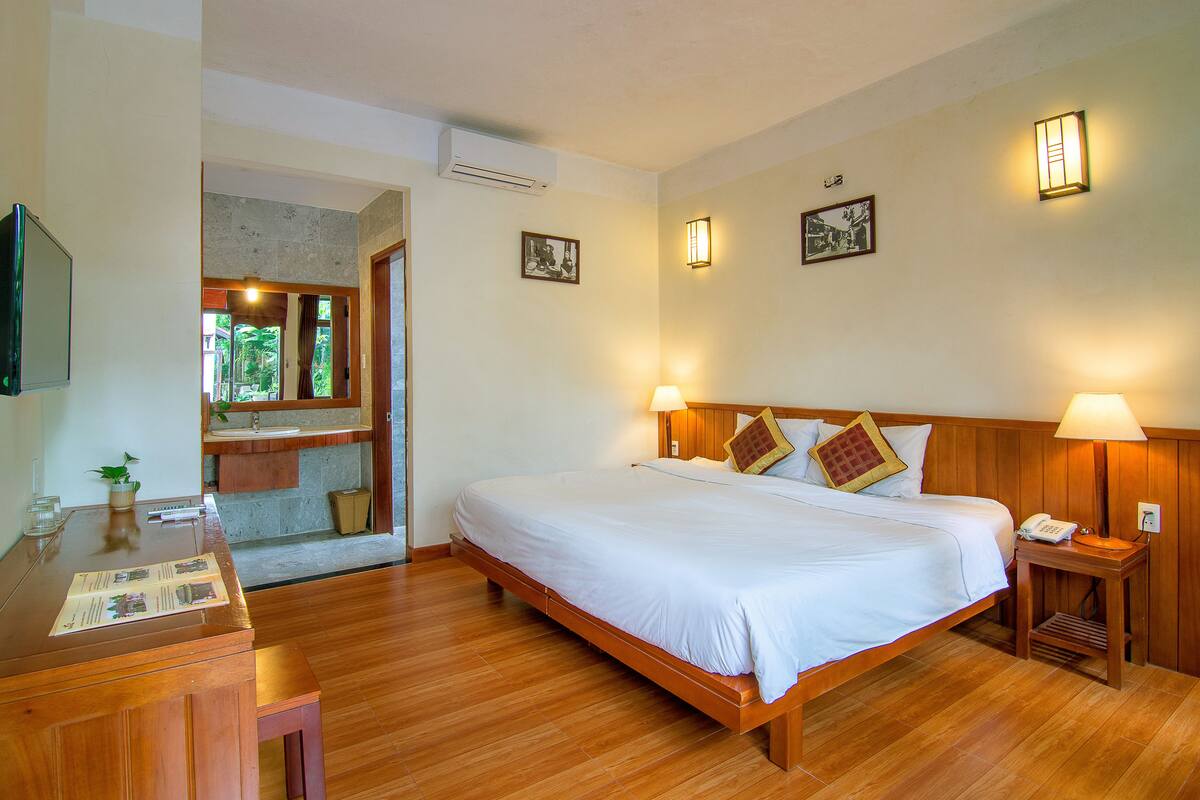 A hotel room with a wooden bed, bedside lamps, a wooden nightstand, a desk with a chair, a mirror, and a small window.