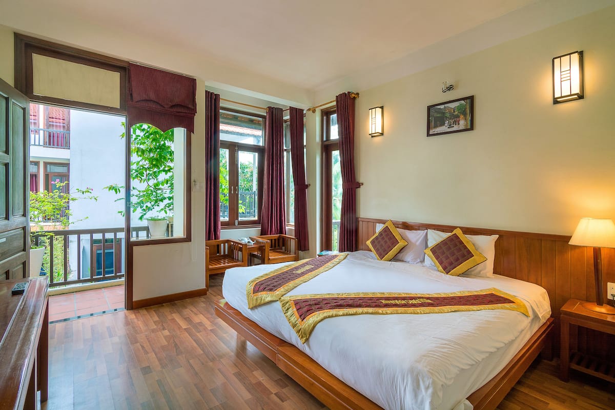 A hotel room with a large bed, wooden headboard, a desk, and a balcony with a view of buildings and greenery.
