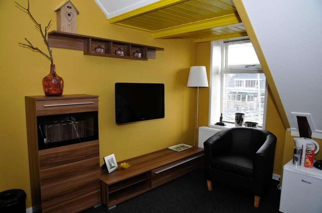 A room with a television mounted on the wall, a black chair, a wooden cabinet with drawers, a small table, and a window with blinds.