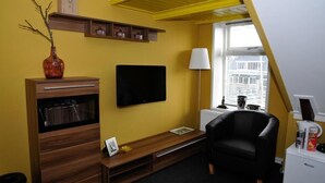 A room with a television mounted on the wall, a black chair, a wooden cabinet with drawers, a small table, and a window with blinds. - Hotel Restaurant It Posthus (Burdaard)