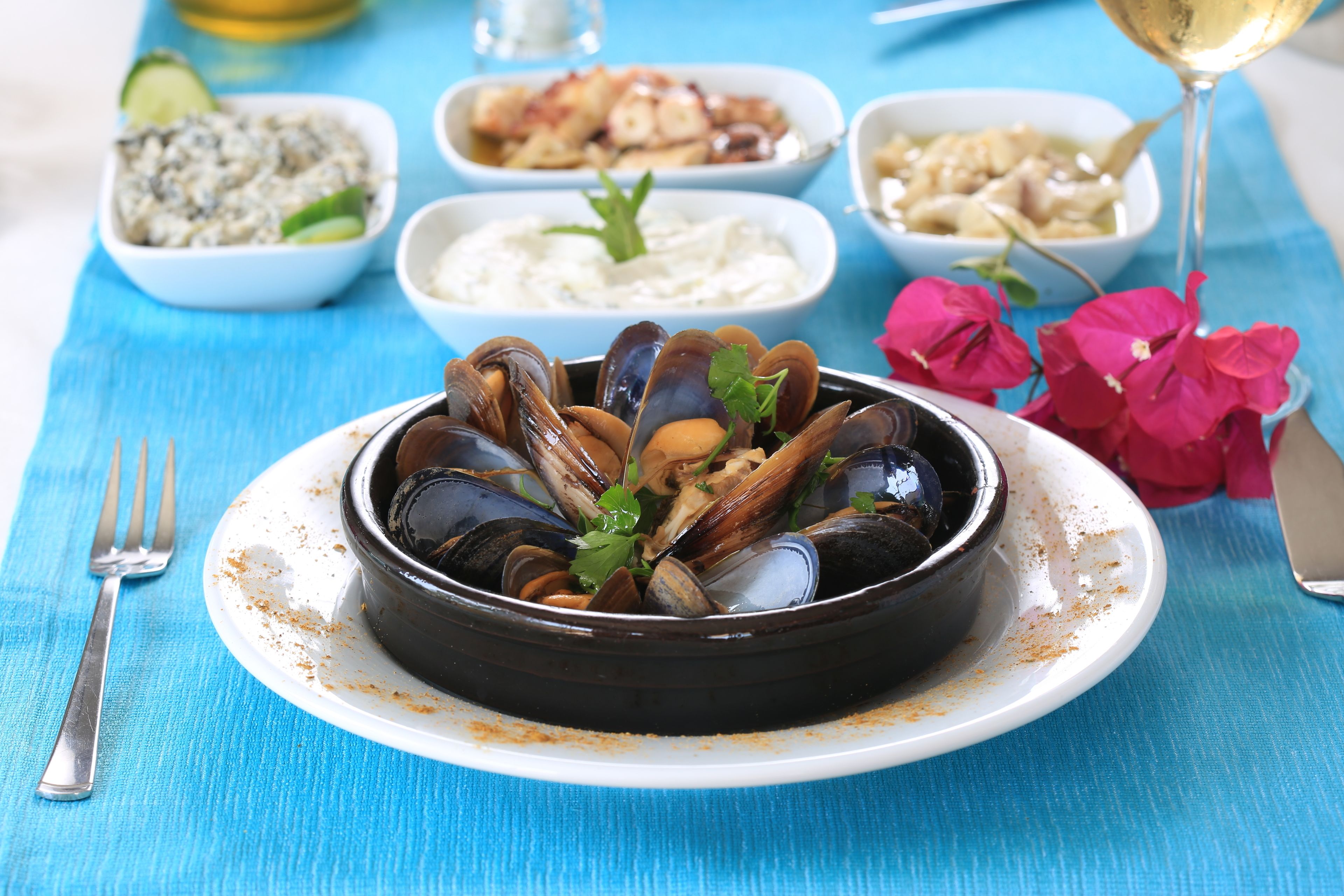 A seafood dish with mussels on a white plate, accompanied by various side dishes and a glass of white wine on a blue tablecloth.
