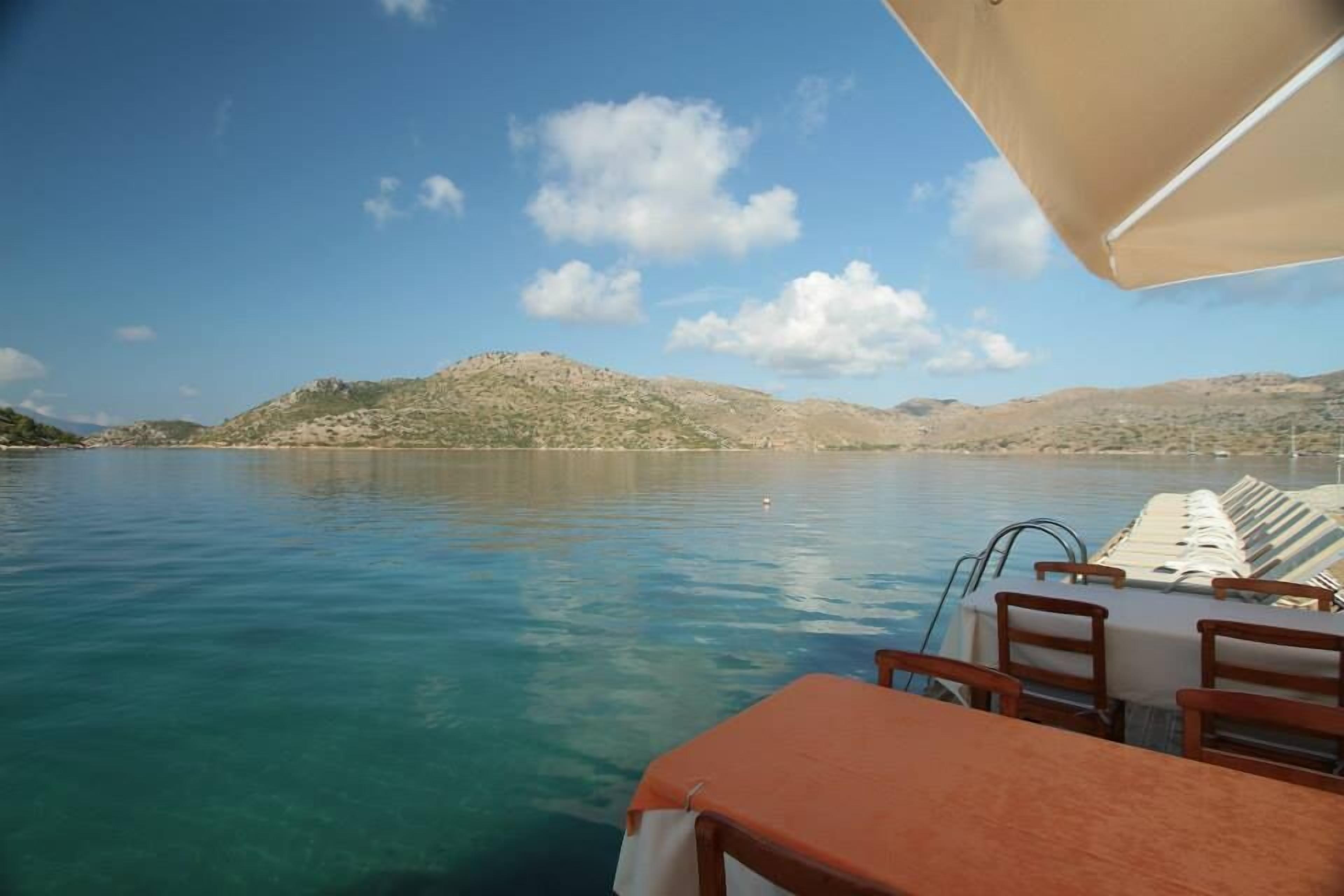 A boat with a table and chairs, a clear blue sea, and distant hills under a blue sky.