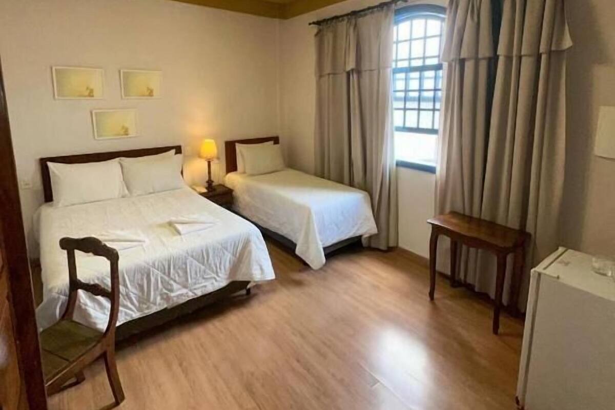 A hotel room with two beds, a small table, a chair, a window with curtains, and a ceiling light fixture.