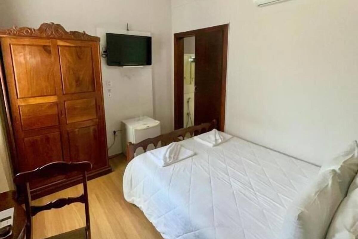 A bedroom with a bed, a wooden wardrobe, a small bedside table, a mirror, and an air conditioning unit on the wall.