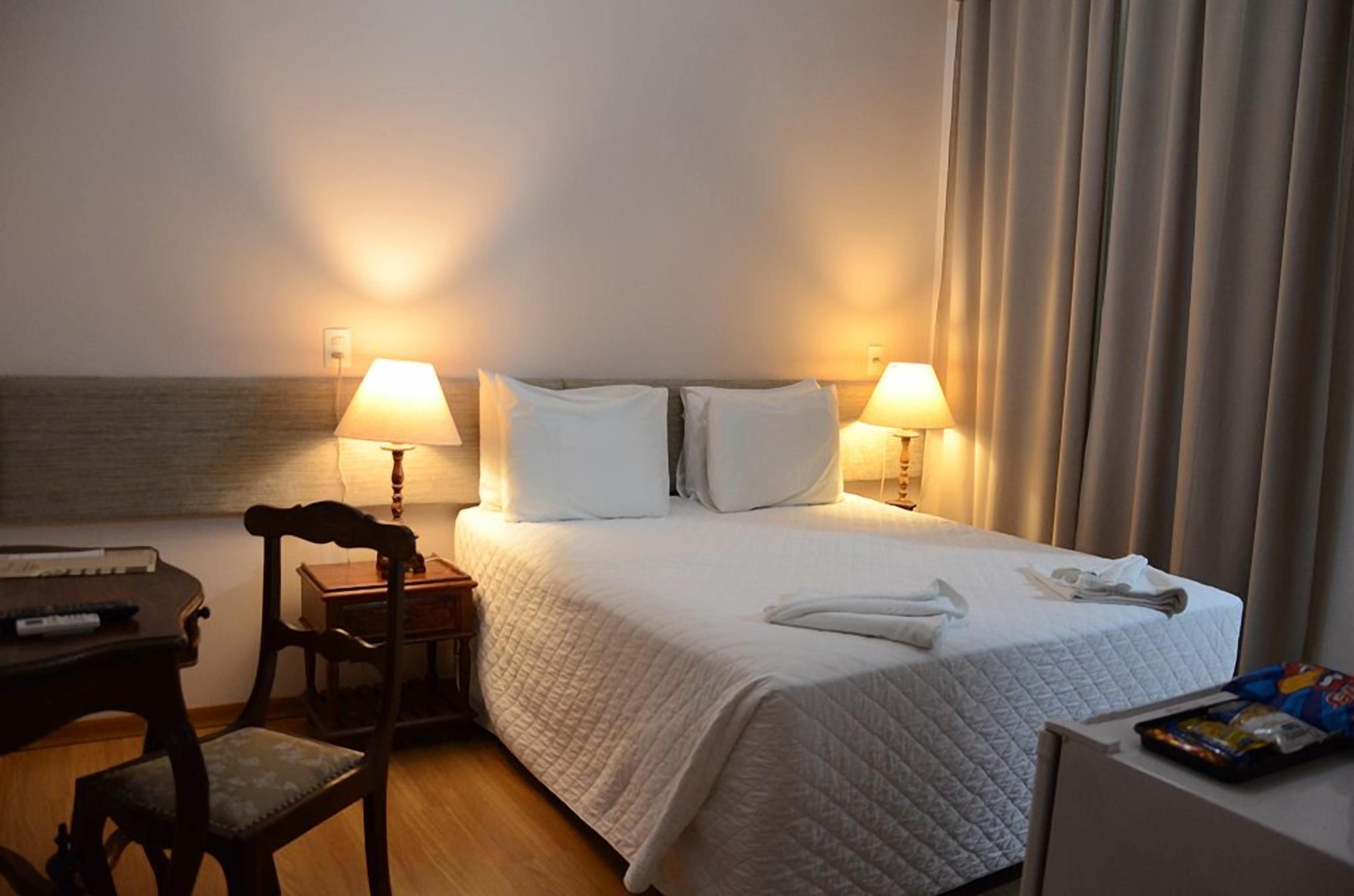 Quarto de hotel com cama, duas luminárias laterais, uma cadeira, escrivaninha e janela com cortinas.