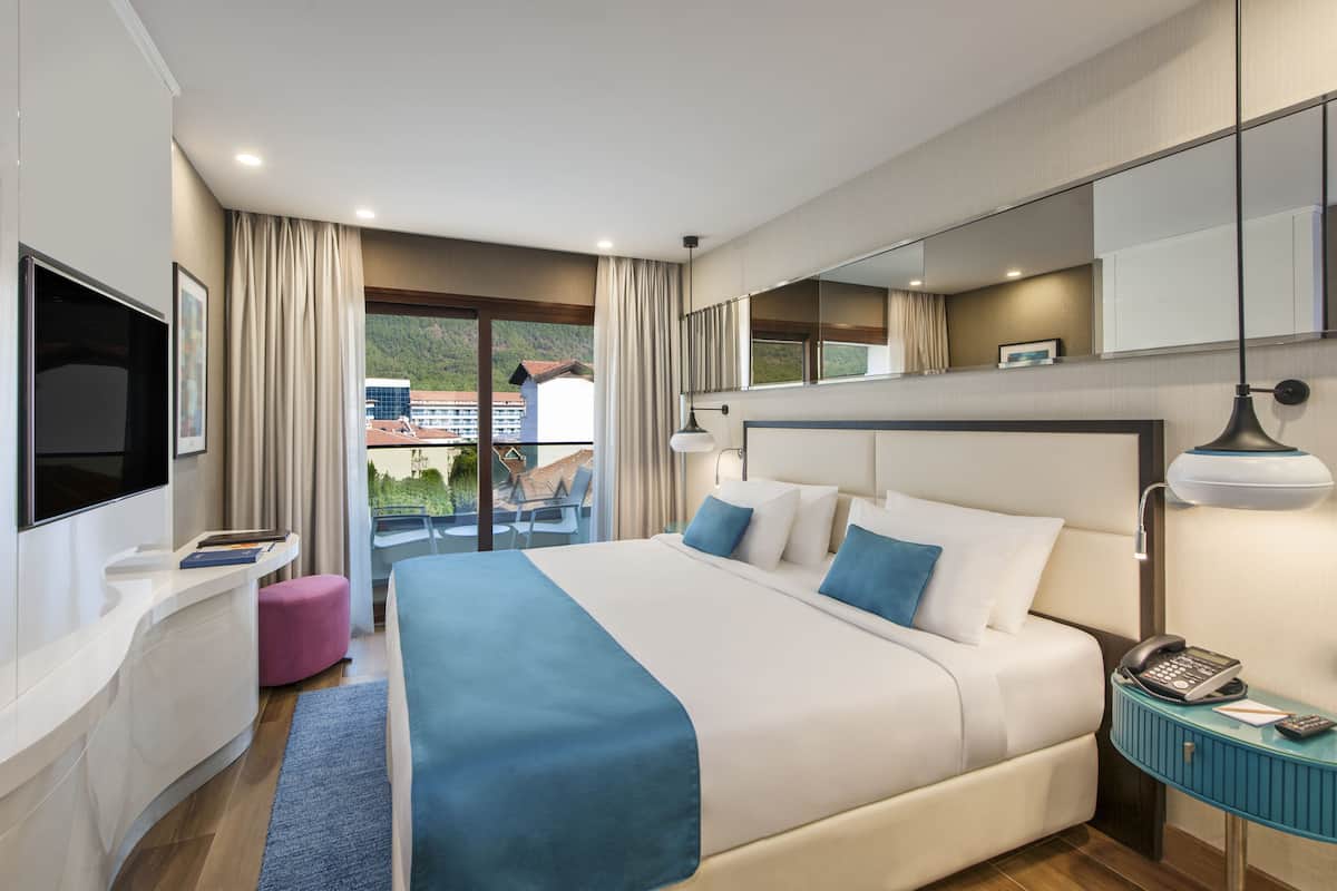 A modern hotel room with a large bed, a desk, a chair, and a balcony with a view of buildings.