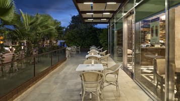 An outdoor patio with tables and chairs, glass walls, and palm trees.