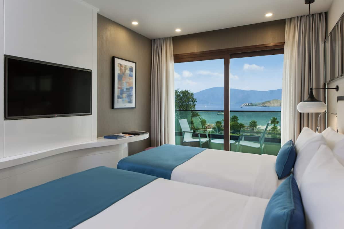 A hotel room with two beds, a TV mounted on the wall, a balcony with a view of the sea and mountains.