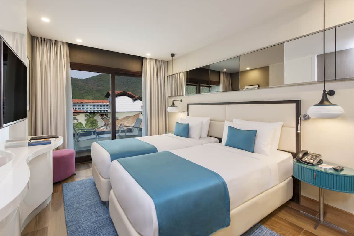 A modern hotel room with two beds, a balcony, a desk, and a phone on a side table.