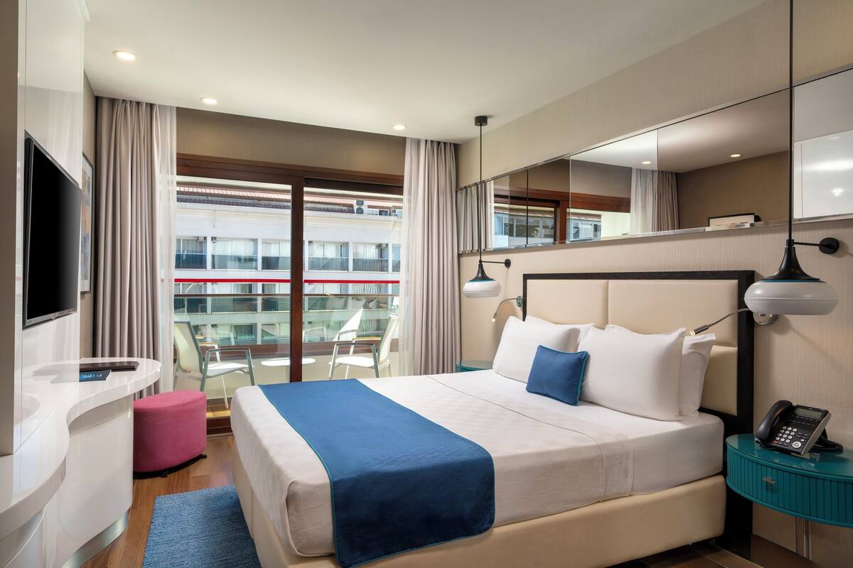 A modern hotel room with a large bed, a desk, a chair, a TV, and a balcony with a view of the city.