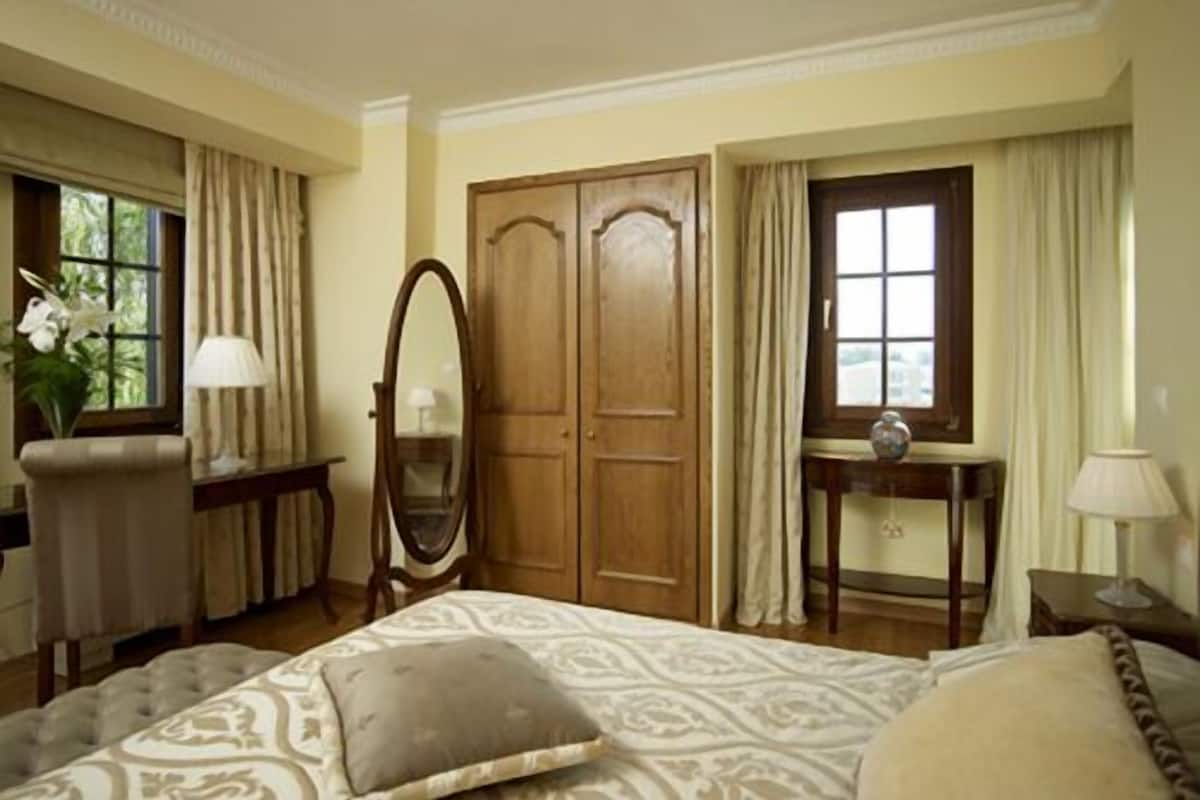 A bedroom with a large bed, a wooden wardrobe, a mirror, and a desk with a lamp.