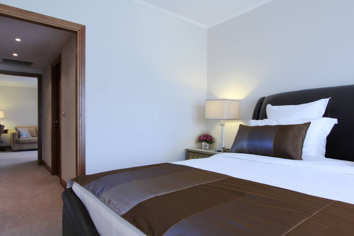A hotel room with a large bed, a bedside table with a lamp, and a view of another room.