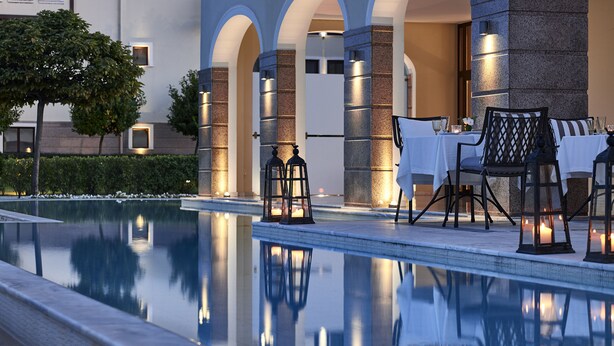 Un área junto a la piscina con mobiliario, una piscina y un edificio con columnas arqueadas y fachada de piedra.