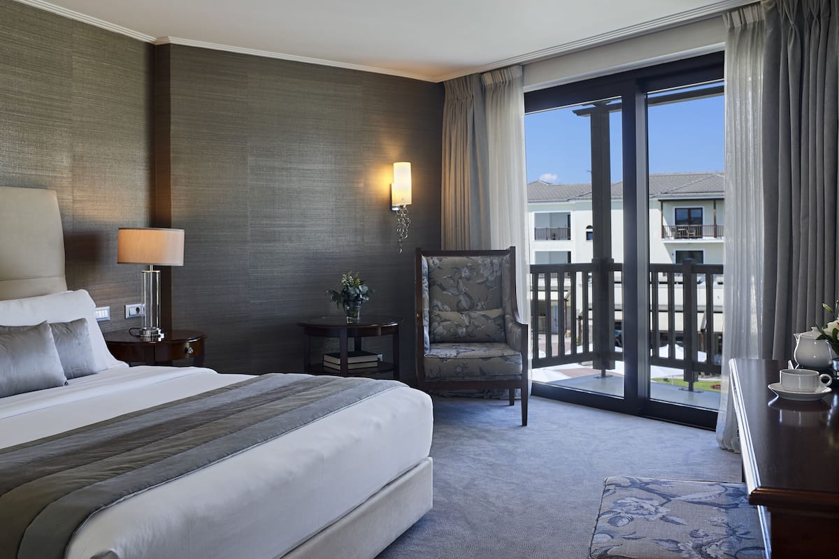 A hotel room with a large bed, a chair, a desk with a cup and saucer, a lamp, a small table with a plant, and a view of the outside through a sliding door.