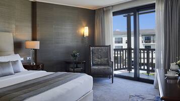 A hotel room with a large bed, a chair, a desk with a cup and saucer, a lamp, a small table with a plant, and a view of the outside through a sliding door.
