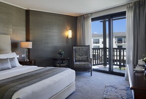 A hotel room with a large bed, a chair, a desk with a cup and saucer, a lamp, a small table with a plant, and a view of the outside through a sliding door. - Hotel Du Lac Congress Center & Spa (Ioannina)