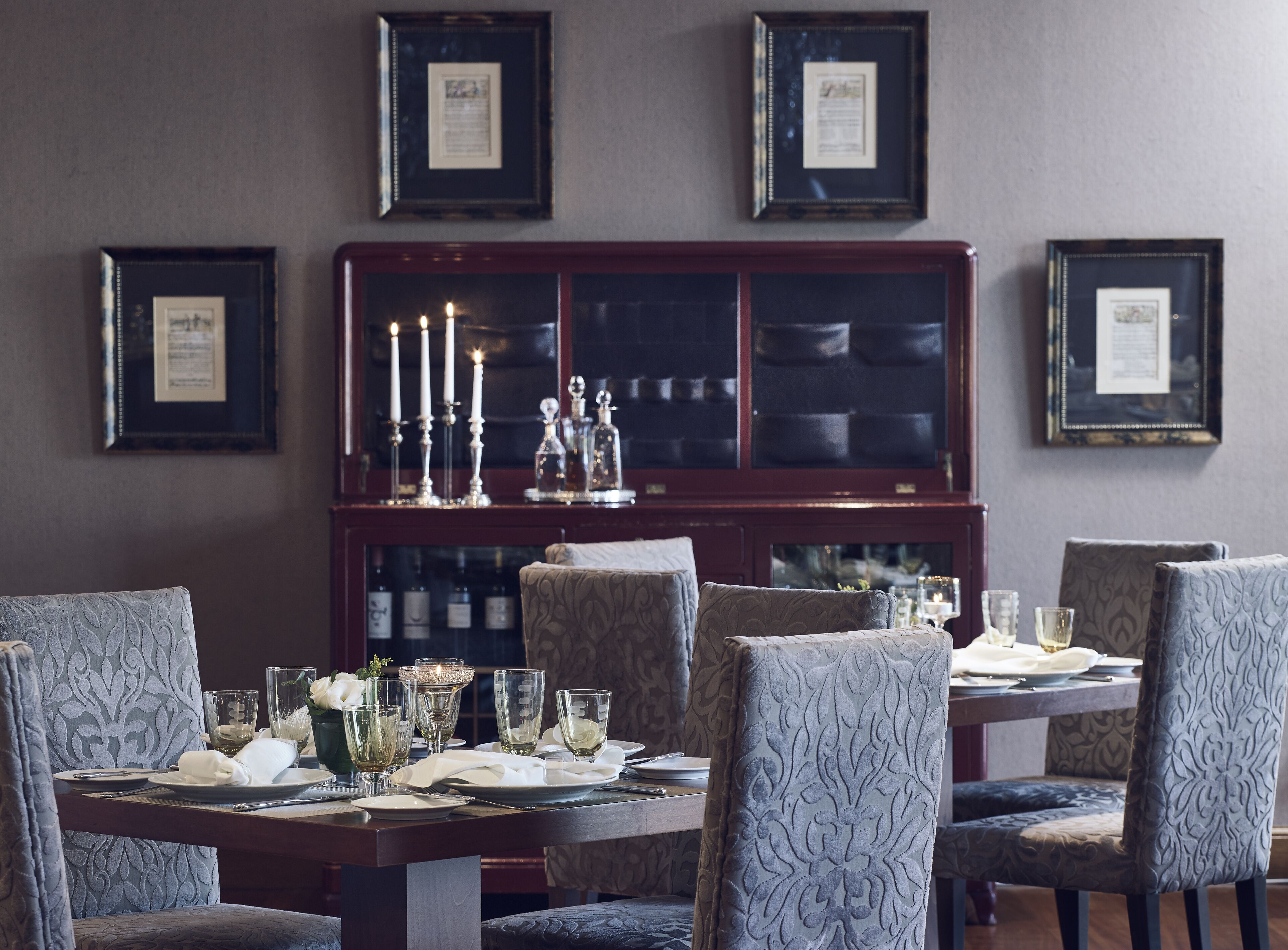 A dining room with a dark wooden cabinet, framed certificates on the wall, and a table set for four with chairs.