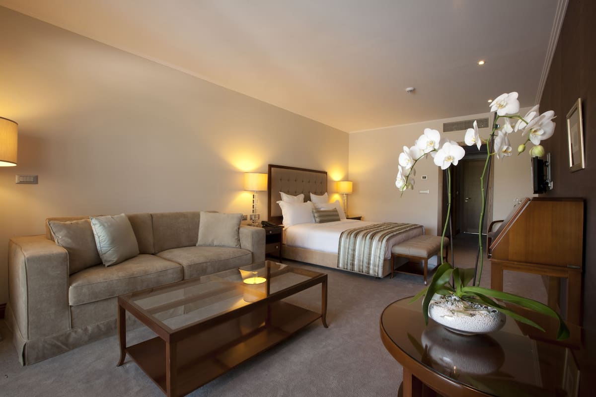 A hotel room with a bed, a sofa, a coffee table, and a dining table with a flower vase.