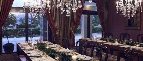 Ein formeller Speisesaal mit langem Tisch, gedeckt für eine Mahlzeit, eleganten Kronleuchtern und Blick auf einen angelegten Garten durch große Fenster.