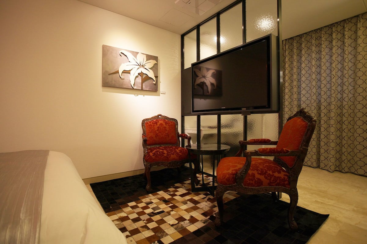 A hotel room with a bed, two ornate chairs, a small table, a flat-screen TV, and a patterned floor.