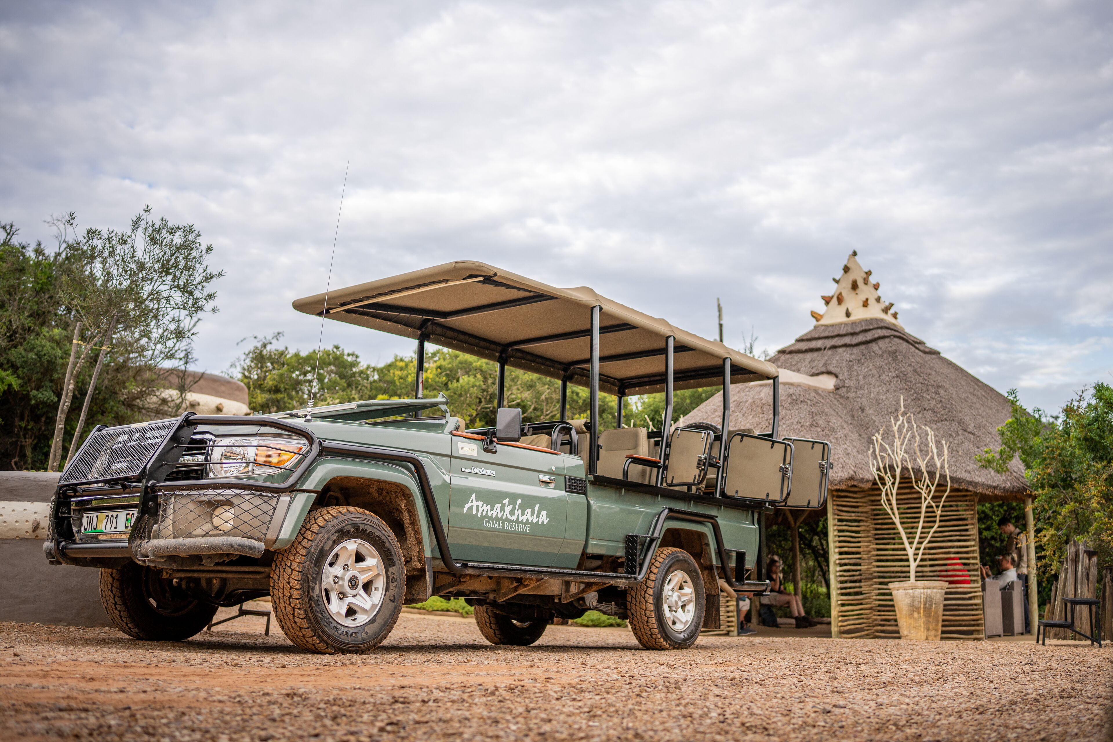 A green SUV with a roof rack parked in front of a thatched-roof structure, possibly a lodge or restaurant, with a gravel path and trees in the background.