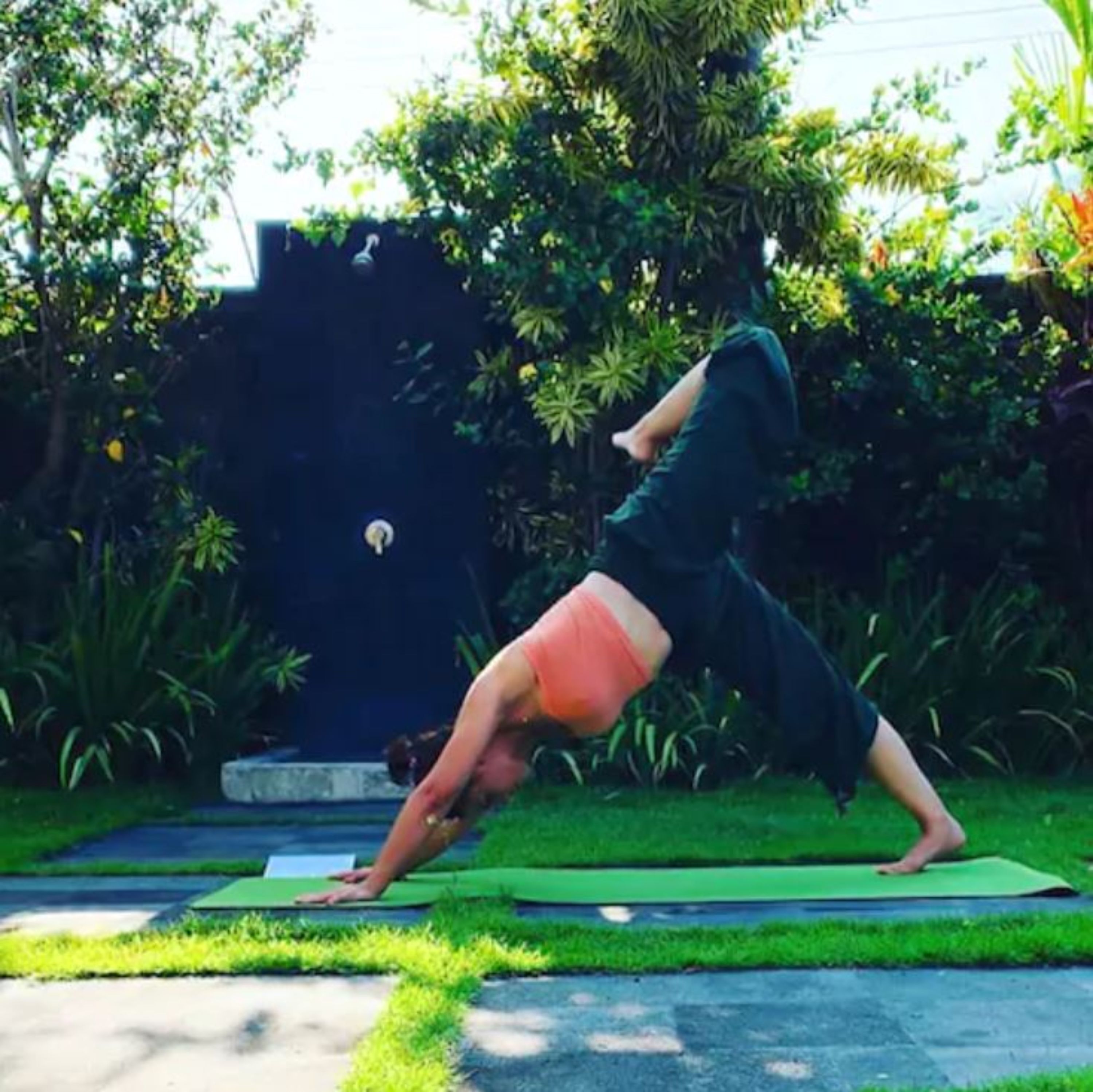 A person performing a yoga pose on a green mat outdoors with trees and a building in the background.