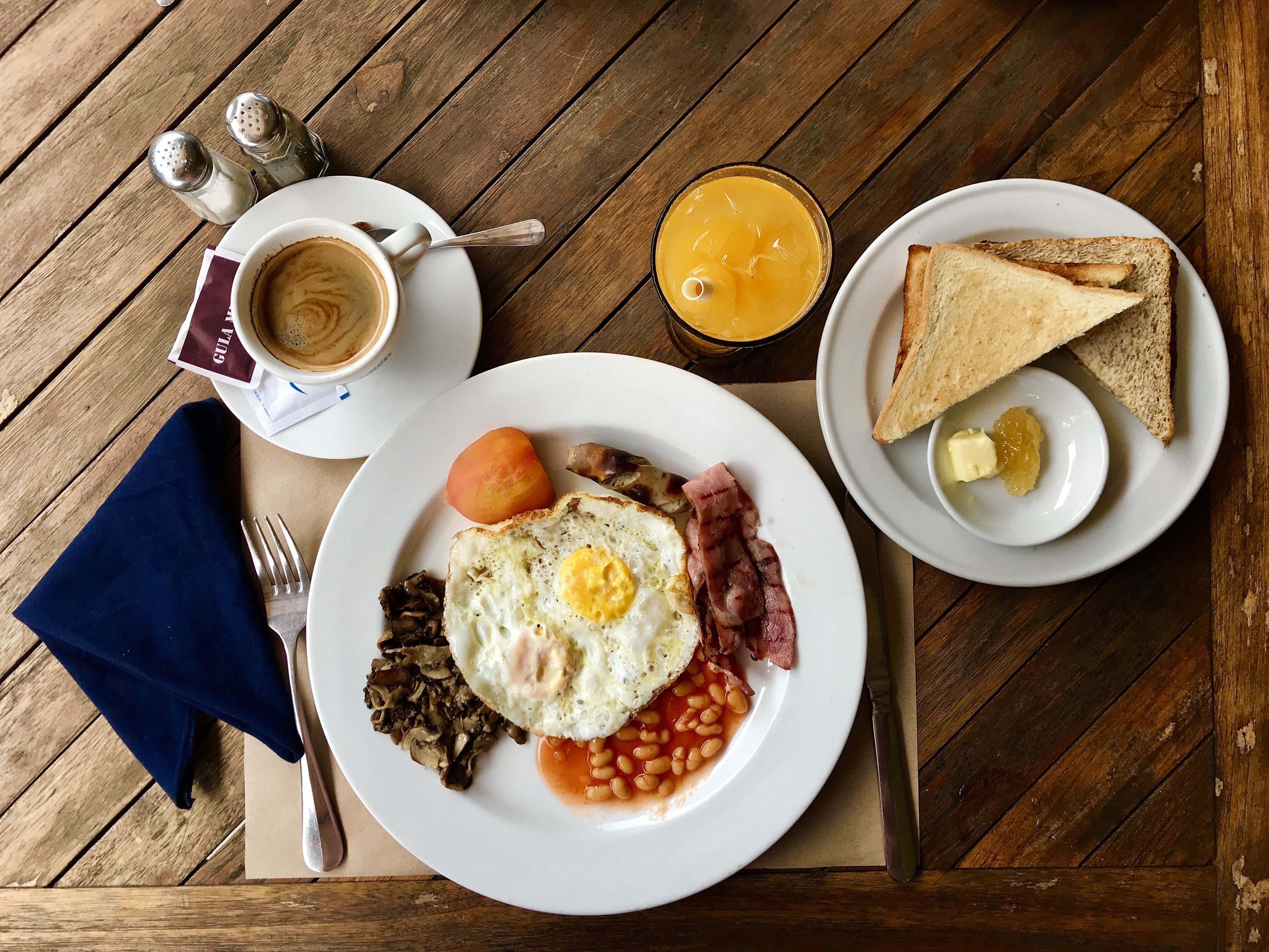 Un plato de desayuno con huevos, tocino, champiñones y frijoles, un vaso de jugo de naranja, tostadas con mantequilla y una taza de café sobre una mesa de madera.