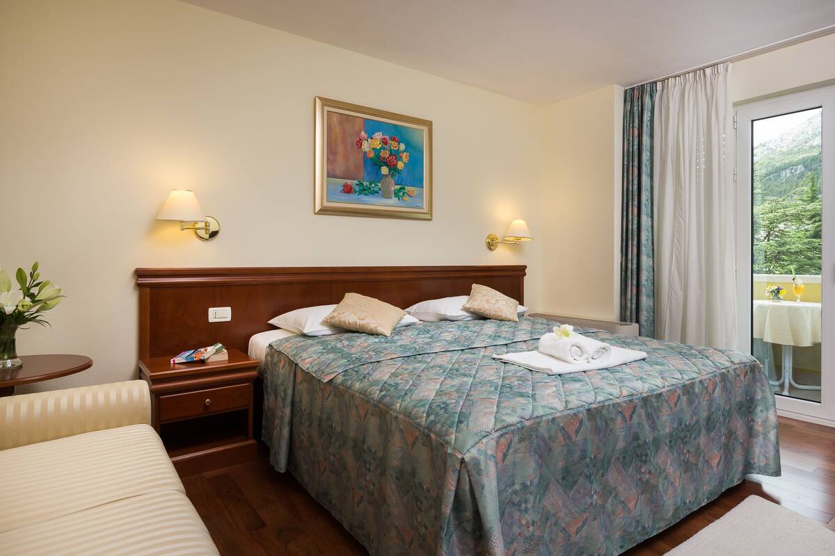 A hotel room with a bed, a nightstand, a bedside table with a vase of flowers, a painting on the wall, and a balcony with a table and chairs.
