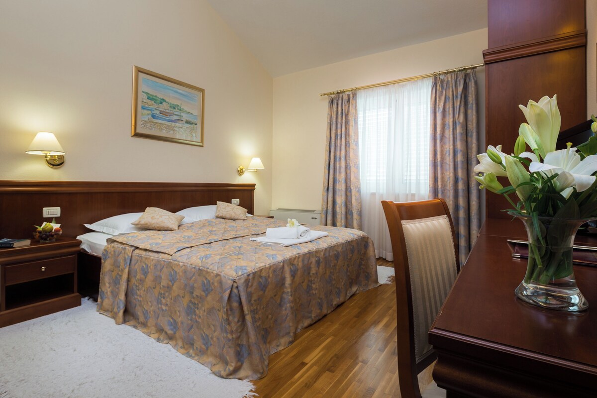 A hotel room with a large bed, two bedside tables, a desk, a chair, a vase of flowers, and a painting on the wall.
