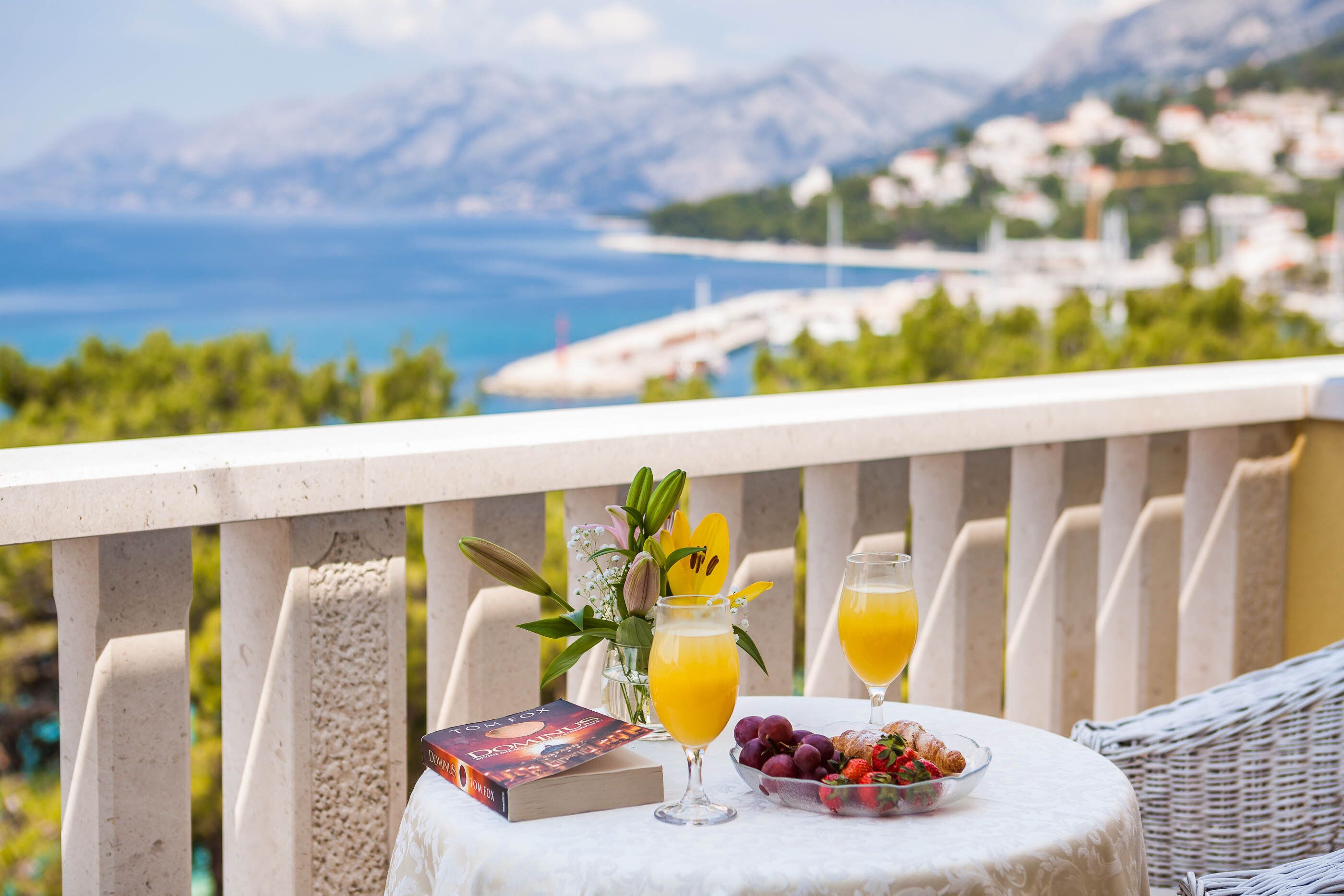 A table with a book, a glass of orange juice, a glass of juice, a bowl of grapes, and a croissant on a balcony overlooking a coastal view.