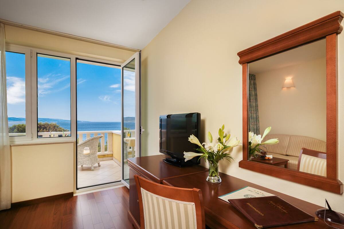 A hotel room with a large window, a television, a desk with a vase of flowers, and a mirror.
