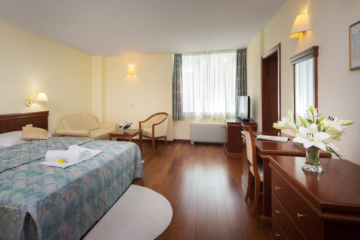 A hotel room with a bed, two chairs, a desk, a TV, and a window with curtains.