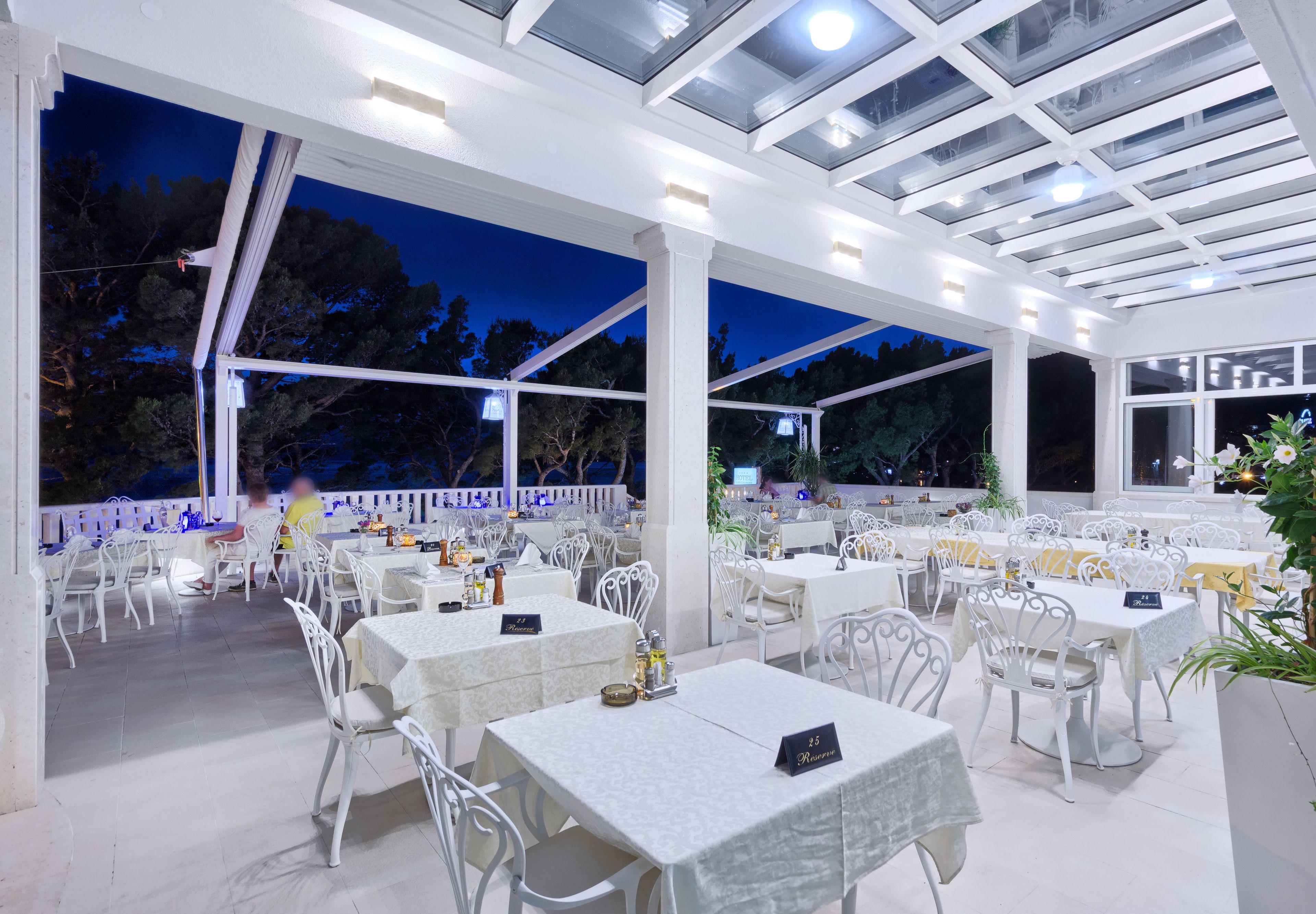 Un restaurante al aire libre con mesas y sillas blancas, cubierto por un techo de cristal, rodeado de árboles.