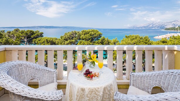 A balcony with wicker furniture, a table with fruit and drinks, and a view of the sea and mountains. - Hotel Bacchus (Baska Voda)