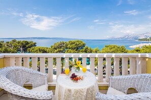 A balcony with wicker furniture, a table with fruit and drinks, and a view of the sea and mountains. - Hotel Bacchus (Baska Voda)