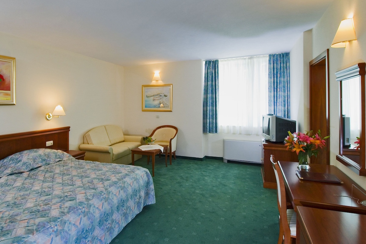 A hotel room with two beds, a chair, a small table, a television, and a painting on the wall.