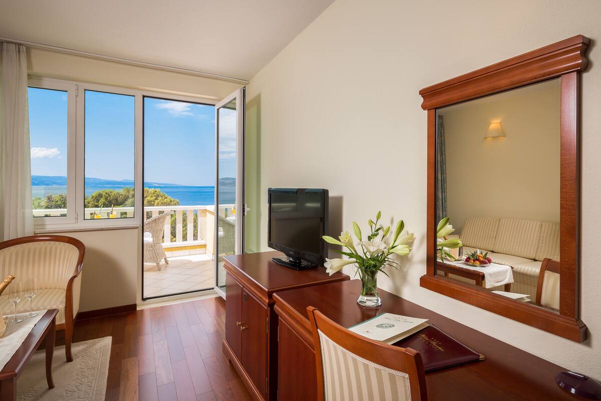 A hotel room with a large window, a television, a desk, a chair, and a mirror.